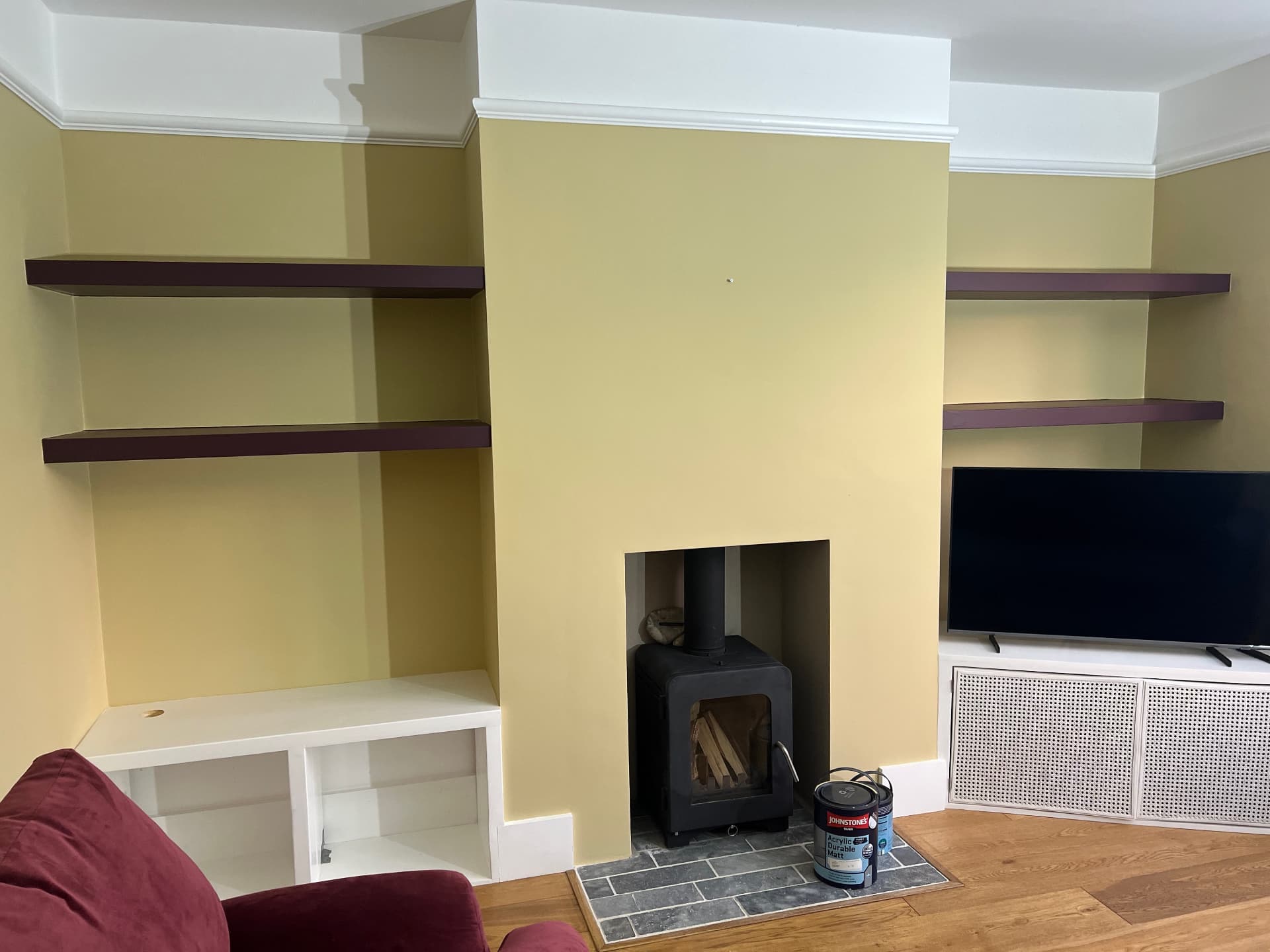 Finished living room with pale yellow walls, deep plum floating shelves and a log burner